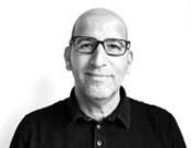 Porträt eines lächelnden Mannes mit Glatze und Brille in einem dunklen Poloshirt vor weißem Hintergrund, schwarz-weiß gehalten.