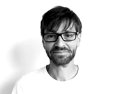 Ein Mann mit Brille und Bart lächelt leicht. Er trägt ein helles T-Shirt. Der Hintergrund ist weiß. Schwarz-Weiß-Fotografie.