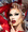 Person mit dramatischem Make-up, roten Lippen, goldenen Ohrringen und Ringen, in rotem Outfit vor metallischem Hintergrund.