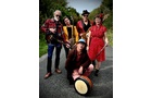 Download Foto: Fünf Personen in bunter, retro-inspirierter Kleidung stehen auf einer Straße, zwei tragen Gitarren, eine spielt eine Bodhrán.