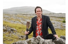 Download Foto: Eine Person steht mit einer Gitarre in einer felsigen, grasbedeckten Landschaft und trägt ein rotes Hemd unter einem dunklen Blazer.