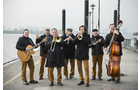 Download Foto: Eine siebenköpfige Band mit Instrumenten steht am nebligen Ufer, darunter Gitarre, Trompeten und Kontrabass, in braunen Hosen.