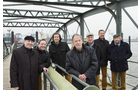 Download Foto: Gruppe von sieben Personen auf einer Metallbrücke, die Kamera lächelnd anblickend. Hintergrund: verschwommene Stadt und Wasser.