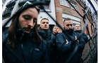 Download Foto: Fünf Männer in dunkler Kleidung stehen hinter einem Maschendrahtzaun und schauen ernst. Gebäude im Hintergrund.