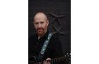 Download Foto: Ein Mann mit rotem Bart hält eine Gitarre vor einer dunklen Wand mit Sterndekorationen. Er trägt ein schwarzes Hemd.