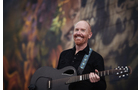 Download Foto: Ein lächelnder Mann mit rotem Bart spielt Gitarre vor einem bunten, unscharfen Hintergrund.