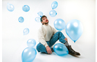 Download Foto: Person sitzt im Studio auf dem Boden, umgeben von schwebenden blauen Luftballons, in legerer Kleidung.