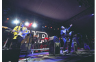Download Foto: Eine Band spielt live auf einer Bühne mit Lichteffekten. Im Hintergrund leuchtet ein großes Logo, Instrumente sind sichtbar.
